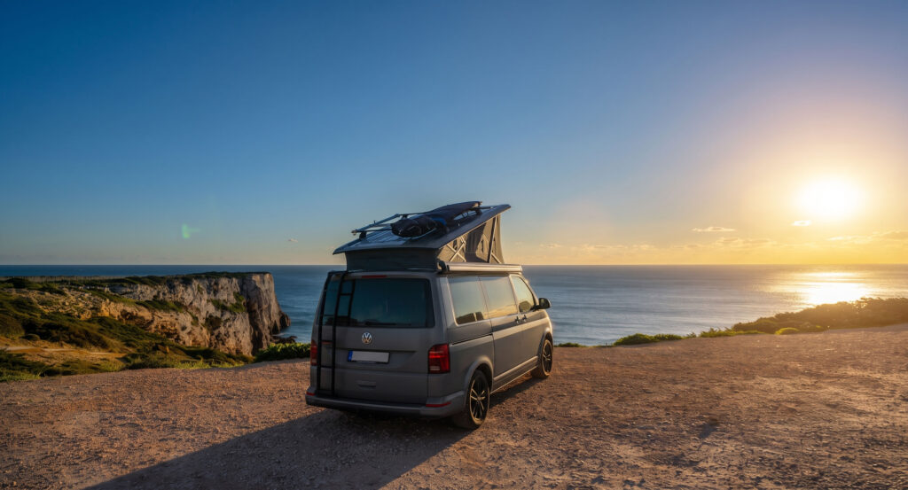 Der VW-Bus mit aufgestelltem Dach und Surfboard steht auf einer Klippe hoch ueber dem Atlantik bei Sonnenuntergang