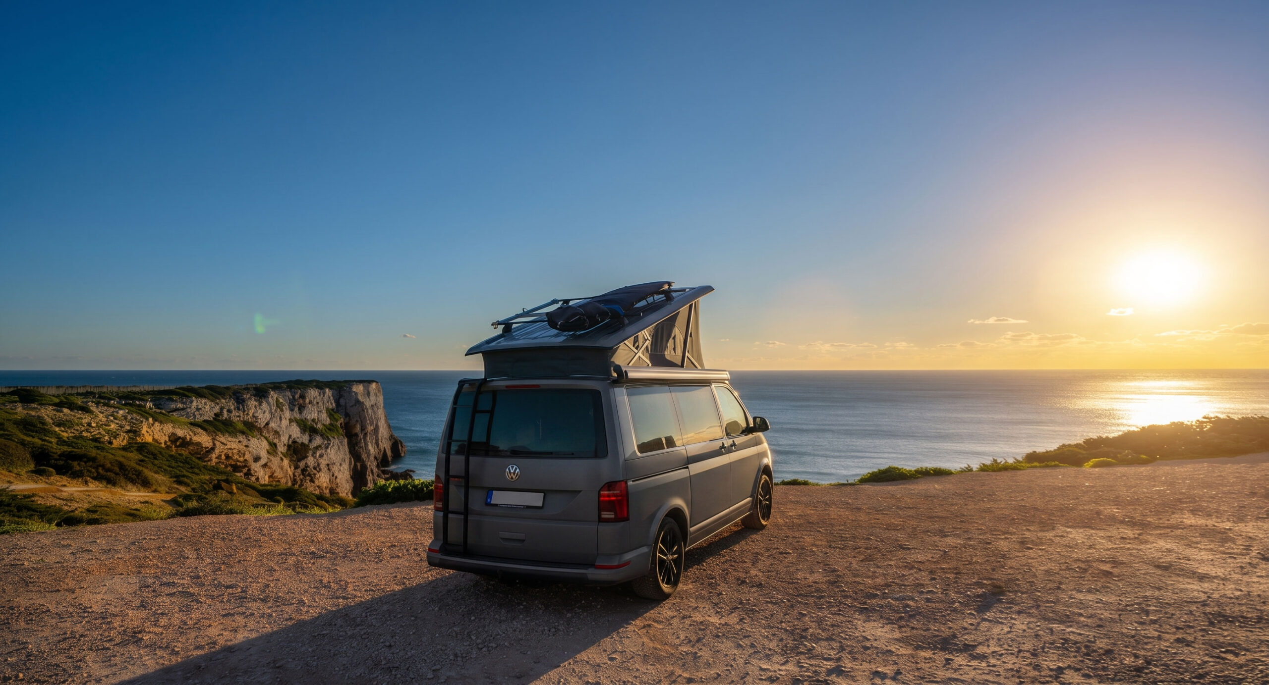 Der VW-Bus mit aufgestelltem Dach und Surfboard steht auf einer Klippe hoch ueber dem Atlantik bei Sonnenuntergang