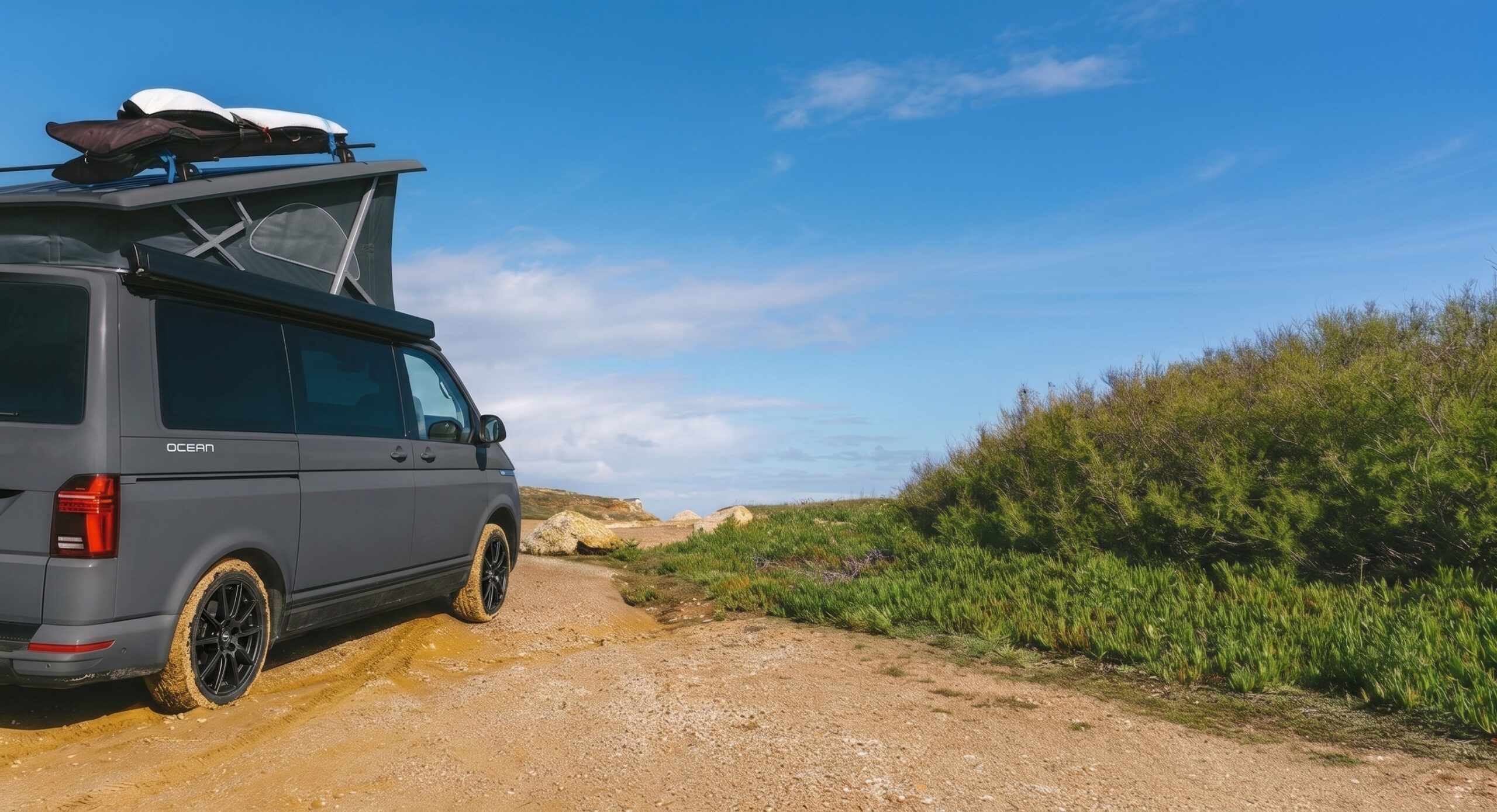 Der VW-Bus mit staubigen Raedern auf einer Sandpiste zwischen Kuestenbewuchs unter blauem Himmel