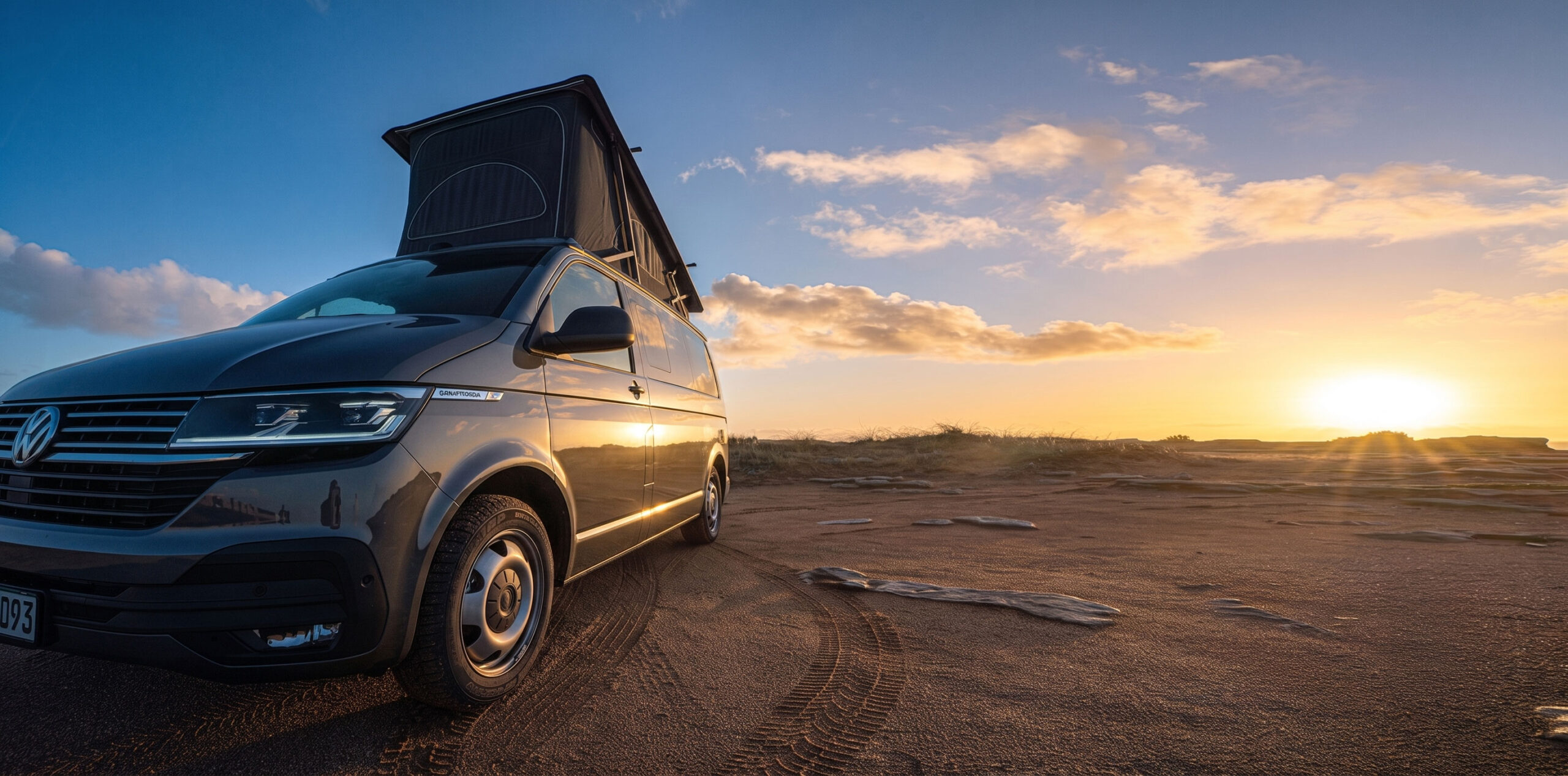 Grauer VW T6.1 California mit aufgestelltem Dach auf nassem Sand, dahinter geht die Sonne auf